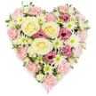 A richly detailed heart-shaped floral tribute is displayed against a bright white background, every flower arranged to form a seamless symbol of love and remembrance often chosen for funerals and memorials in Stanmore. The heart is densely packed with blossoms so that it appears plush and three-dimensional, almost like a soft floral cushion. At its centre, several prominent roses in a creamy, pale yellow take pride of place, their layered petals curving inward to form classic spiral rosettes that suggest warmth, gratitude, and enduring affection. Radiating out from these central blooms, an abundance of pastel pink carnations fills most of the heart, their delicately fringed petals overlapping to create a gently textured surface that defines the soft contours of the heart's outline. Nestled among the carnations are bright white spider chrysanthemums, recognisable by their fine, needle-like petals fanning out from greenish centres, adding brightness and an intricate, almost feathery contrast. Deeper magenta-pink lisianthus blooms are scattered thoughtfully throughout, their rounded, cup-like shapes and rich colour adding emotional depth and drawing the eye across the composition. Small hints of lush green foliage and tiny buds weave between the flowers, suggesting freshness and the care of expert hands. At the base point of the heart, a pale pink ribbon peeks out, hinting at a dedication or message of condolence that might accompany the tribute when delivered. The overall mood of the arrangement is one of quiet dignity and heartfelt compassion, a floral heart that gently honours a life and offers comfort to those mourning a loss.