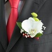 A close-up photograph shows a wedding boutonniere pinned neatly to the lapel of a dark charcoal grey suit jacket, the subtle woven texture of the fabric catching soft indoor light. At the heart of the design sits a single, flawless white Phalaenopsis orchid, its broad, velvety petals opening like wings around a yellow and white lip that draws the eye. Tucked just above the orchid are two small, round lime-green chrysanthemum-style blooms, their tight, cushion-like petals adding a playful pop of colour against the formal suit. Fine sprays of white baby's breath weave delicately around the main flower, each tiny blossom like floating mist, softening the overall silhouette. A few glossy, rich green leaves cradle the orchid from behind, giving structure and a natural frame to the arrangement. The stems are bound with a narrow, deep red satin ribbon that mirrors the nearby solid red satin tie, which stands out boldly against a pale grey and white checked dress shirt. Light glides over the smooth orchid petals, the slight sheen of the ribbon, and the crisp lapel edge, highlighting the thought and craftsmanship in each placement. The boutonniere sits just above a neatly stitched jacket pocket, perfectly positioned to be noticed during a ceremony or reception. The mood is elegant and modern, the kind of refined detail you might see on a groom walking through Stanmore before heading to a wedding at a local church or a reception near Bentley Priory, carrying with him this small but meaningful piece of floral artistry.