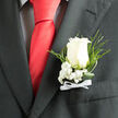 In this close, intimate view, a classic white rose boutonniere is pinned neatly to the lapel of a dark charcoal suit, capturing the graceful balance between formality and emotion that so often defines celebrations in Stanmore. The central rose is an ivory white, gently opening with layered petals that spiral outward with effortless elegance. Each petal has a soft, velvety texture, and the light picks up subtle shadows in the folds, giving the bloom a three-dimensional presence, as if you could almost reach out and touch it. Cradling the base of the rose are clusters of baby's breath, their numerous tiny, round blossoms creating a misty, cloudlike effect that frames the rose without overwhelming it. This delicate white haze softens the visual contrast between flower and suit, just as a slight morning mist might soften the skyline over Stanmore Hill. Nestled behind the main flower, slender green ferns and looping strands of glossy foliage bend and curve with gentle movement, introducing a natural, organic rhythm to the design. The stems are bound with care, and a neat white ribbon bow sits at the base, its satin finish catching a restrained sheen of light where it meets the pocket edge. The overall look is measured and refined, yet undeniably romantic, echoing the atmosphere of a ceremony held at a local church or register office before heading to a reception near Canons Park. You can almost sense the quiet confidence and pride of the wearer-perhaps a groom or prom guest-standing tall as they greet family and friends, the boutonniere serving as a small emblem of promise and celebration.