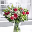 A richly detailed photograph shows a romantic red and white bouquet arranged in a classic clear glass vase, resting on a bright white tabletop near a softly sunlit window in Stanmore. At the heart of the arrangement, velvety crimson roses in various stages of bloom draw the eye: some roses are fully open, revealing layered, plush petals, while others remain in tight, elegant buds that promise to unfurl over the coming days. Nestled around them are ruffled red carnations, their densely packed petals adding volume and a slightly old-fashioned charm that many families along The Broadway still associate with traditional celebrations. Threaded between these deep red blooms are pristine white alstroemeria, also known as Peruvian lilies. Some alstroemeria flowers are open just enough to reveal their slender, trumpet-shaped petals tipped with a hint of green, while others are still closed buds, like little torches of future colour. Delicate sprays of tiny white gypsophila, or baby's breath, form a soft mist around the bolder blooms, catching the gentle daylight that filters through the nearby window and creating an almost cloud-like halo. Lush greenery forms a strong base to the design: broad, glossy deep green leaves encircle the stems at the base, giving structure and framing the arrangement, while more slender, lighter green foliage weaves upward, adding movement and texture. Through the sides of the cylindrical glass vase, the neatly trimmed stems are clearly visible, immersed in clean water that suggests just-delivered freshness, as though this bouquet has only just arrived from a local florist near Stanmore Hill. Behind the flowers, the background is quietly blurred in soft grey tones, making the vivid reds, whites, and greens stand out with striking clarity. The overall impression is one of warm appreciation and joyful celebration, suitable for birthdays, anniversaries, heartfelt thank-yous, or any moment when someone in Stanmore deserves a beautiful, uplifting surprise delivered to their door.