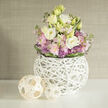 Bathed in soft morning light, this elegant round bouquet pairs frothy creamy-white and pale yellow lisianthus with gentle clusters of blush-pink stock and small, architectural purple allium heads, all woven into a lush embrace of fresh green foliage. The flowers are housed in a distinctive sculptural vessel: a white, spherical lattice of slender interlaced twigs that lets stems peek through and lends a contemporary, airy silhouette. To the left, two hand-woven natural wood spheres rest on the clean white tabletop, mirroring the vase's woven pattern and tying the display together with a relaxed, artisanal warmth. Behind the arrangement a neutral, textured greige wall-resembling linen or grasscloth-provides a serene backdrop that allows the pastel palette to softly radiate.
As a Stanmore florist familiar with local homes and the light by Canons Park and Bentley Priory, I imagine this composition brightening a compact dining nook or sitting room near Stanmore tube station. The scent would be gentle-soft floral musk with a fresh green lift-and the bouquet's tactile story is evident in every element: the velvet-soft lisianthus, the feathery stocks, the spiky punctuation of allium and the cool, smooth white finish of the woven vessel. Flower Delivery Stanmore prepares pieces like this for understated celebrations, get-well gestures, or to comfort and soothe; it feels crafted for neighbours who appreciate contemporary form married to natural textures and local, thoughtful styling.