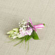 Placed gently on a neutral, subtly woven cloth that gives the background a soft, homely texture, this refined boutonnière embodies the careful craft of a Stanmore wedding florist. At the centre sit creamy buds, likely freesia, their petals firm yet slightly translucent, as if holding the light that falls from a nearby window. They are paired with clusters of pale pink flowers whose petals curve outward with a whisper-soft ruffle, the blush tone reminiscent of the evening sky over Stanmore Country Park. Airy sprays of gypsophila-baby's breath-hover between and above the main blooms, their tiny white florets forming a delicate mist that softens every edge. Slender, grass-like strands arc gracefully through the arrangement, adding a gentle sense of movement, while broader, deep-green leaves curl protectively around the base, giving structure and depth. The stems are wrapped in a smooth, satin ribbon of rose-pink, carefully wound and finished with a perfectly tied knot whose slight sheen catches the light. You can almost sense the fragrance: a soft, fresh blend of floral and green notes, the kind of gentle scent that lingers close to the wearer without ever becoming overpowering. This small floral piece feels deeply personal, like something chosen thoughtfully for a groom getting ready in a Stanmore townhouse or for a prom date posing on a quiet street off Church Road. Crafted in the spirit of local celebrations and family gatherings, the boutonnière adds a subtle brush of colour and natural grace to a lapel, reflecting the understated elegance and warm sentiment so often seen in Stanmore's special occasions.