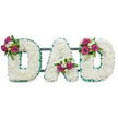 A dignified three-dimensional tribute spells the word DAD in large, capital letters, each letter formed from a dense carpet of pristine white chrysanthemums that create a soft, cushion-like surface against a pure white backdrop. The tiny blooms are packed closely so the letters read as cloud-white blocks, their rounded petals catching a cool, diffuse light that suggests a quiet morning in Stanmore. A ruffled, emerald-green ribbon traces both the outer and inner edges of each character, its pleated texture adding crisp definition and a steady, respectful contrast to the white mass of flowers. Behind the letters a subtle dark green or grey horizontal bar supports the piece, hinting at the solid easel or stand used for graveside or chapel display.
Carefully balanced accent clusters punctuate each letter: the first D carries a spray of rich magenta roses and small white freesias on its upper left curve; the A has its cluster nestled gracefully in the lower V; the final D mirrors the first with a matching arrangement on its upper right. The roses offer a warm, velvety note while the freesia brings a delicate, honeyed scent, and deep green foliage gives depth and structure. The overall feeling is one of reverent tenderness-traditional and personal-a floral voice for goodbye. A local florist from Stanmore might imagine this placed beside a polished pew at St. John's or at a graveside near Stanmore Common, where the clean colours and quiet lines feel at once respectful and intimately consoling.