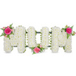 This image shows a beautifully composed funeral flower arrangement spelling the word MOM in clean, capital letters, resting against a spotless white background so the colours and shapes stand out in gentle relief. Each letter is entirely formed from small white flowers, most likely spray chrysanthemums, placed closely together to create a dense, padded texture reminiscent of a soft cushion or carefully tended lawn. The bright white petals convey purity, respect, and stillness, echoing the quiet atmosphere of memorial spaces around Stanmore. Supporting the floral letters, a dark green framework or band is visible along their bases, providing subtle contrast and the reassurance of sturdy construction for display at a service or burial. To lift the tribute from purely white into something more personal and affectionate, three clusters of pink roses and green foliage are integrated into the design. The first nestles at the top left of the opening M, featuring a striking fuchsia rose surrounded by lighter pink roses and buds, with delicate fern fronds trailing outward. A second cluster is placed slightly lower, between the O and final M, giving the impression of the word being gently held from beneath. The third cluster crowns the upper right of the last M, completing a diagonal line of colour that leads the viewer's eye across the entire piece. The overall feeling is one of thoughtful craftsmanship and quiet devotion, the kind of floral tribute that might be arranged by Flower Delivery Stanmore for a family wishing to honour their mother's life with a design that is both traditional and deeply heartfelt.