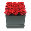 An immaculate arrangement of twelve preserved deep red roses is presented in a sleek square black gift box, the sort of luxurious piece you might have delivered to a modern home in Stanmore for an anniversary or Valentine's celebration. Each rose is fully open, its petals layered in lush, velvety spirals that create a dense, textured surface of crimson, with subtle shifts in tone from dark ruby in the shadows to a slightly brighter red where the soft light touches the petal edges. The blooms are arranged in three neat rows of four, packed tightly so that the top of the box is entirely filled, with the rose heads lifting just above the rim for a generous, overflowing appearance. The box itself is fashioned from matte black material, with clean, sharp corners and a smooth, unembellished surface that gives a sense of modern minimalism. A slender white stripe encircles the lower part of the box like an understated trim, providing a polished, contemporary contrast. The background is a pure, uninterrupted white, isolating the arrangement and making the colour contrast between the roses and the box even more striking, much like a statement piece on a glass table in a Stanmore Broadway apartment or a chic home near Canons Drive. Soft, balanced lighting creates gentle, natural shadows between the petals and along the box edges, enhancing the three-dimensional quality without introducing any harsh reflections. The overall feeling is of refined romance and enduring luxury, perfectly suited for heartfelt gestures and intimate celebrations across Stanmore and nearby neighbourhoods.