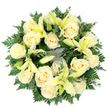 Viewed from directly above on a pristine white background, this circular sympathy wreath reads as a careful study in calm: a densely packed ring of soft cream roses and pale, lemon-yellow lilies cradled by deep green foliage. Around fifteen fully opened roses, their petals tightly spiralled in classic ivory tones, create the wreath's luminous backbone while several gentle lilies-some wide-open with trumpet-shaped petals and pale yellow stamens, others in bud or half-revealed-introduce a slender, graceful height and a whisper of citrus-tinged scent. The flowers sit on a rich bed of mixed greens: feathery fern fronds that give a soft, lacy edge and clusters of small, rounded leaves-likely eucalyptus or similar-that add cool silvery undertones and a firmer texture. Soft daylight seems to lift the cream and lemon hues against the green, the composition balanced and symmetrical so the circular silhouette reads as an unbroken token of remembrance. The arrangement feels tactile as well as visual; the velvety rose petals, the satiny lily curves, and the airy fronds combine to create a comforting, layered touch. Speaking as a local florist who often prepares tributes for services in Stanmore and gatherings near Stanmore Common, I picture this wreath laid gently at a graveside or on a chapel table, offering a quiet message of peace, purity, and consolation. Its restrained palette and refined craftsmanship make it a dignified choice for a funeral, memorial, or private moment of remembrance in the Stanmore community.